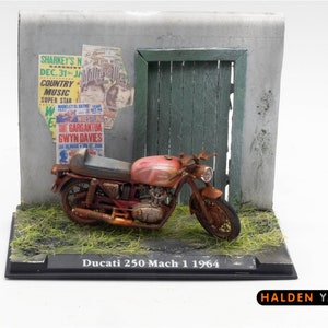 Peut inclure: Modèle de moto Ducati 250 Mach 1 de 1964 de couleur rouge rouillée, garée devant un mur gris délabré avec une porte en bois verte. La moto est sur un socle noir avec le texte "Ducati 250 Mach 1 1964" et "HALDEN YARD" imprimé dessus.