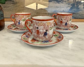 May include: Three white porcelain teacups with red and gold trim and a floral pattern featuring women in traditional Japanese clothing. The cups are on matching saucers with a similar pattern.