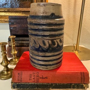May include: A vintage stoneware pottery vase with blue and brown stripes and a wavy design. The vase is sitting on top of a stack of books with the title "The Last Hero" visible.