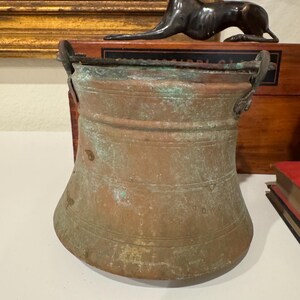 May include: An antique copper bucket with two handles. The bucket has a patina and is slightly dented.