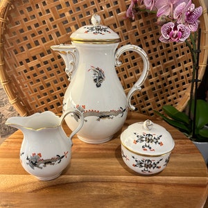 May include: A white porcelain tea set with a black and orange floral design. The set includes a teapot, creamer, and sugar bowl.