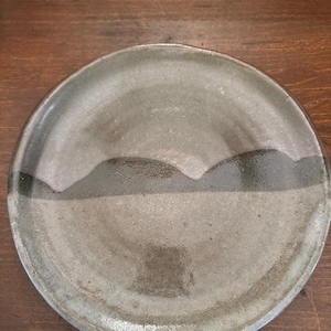 May include: A round ceramic plate with a light gray glaze and a dark gray band around the edge. The band has a wavy, mountain-like design.
