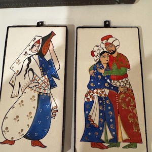 Puede incluir: Dos azulejos de cerámica con escenas pintadas a mano de personas con vestimenta tradicional turca. El primer azulejo representa a una mujer con un vestido blanco, azul y dorado sosteniendo una jarra. El segundo azulejo muestra a una pareja con ropa colorida abrazándose.