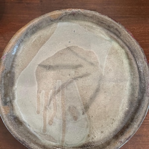 May include: A round ceramic plate with a light brown glaze and a darker brown rim. The glaze has a unique pattern of drips and swirls, creating a rustic and organic look.