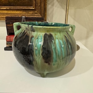 May include: A round, glazed ceramic planter with a bulbous shape. The planter features a dark green and black color scheme with a dripped glaze effect. It has two handles and three small feet.