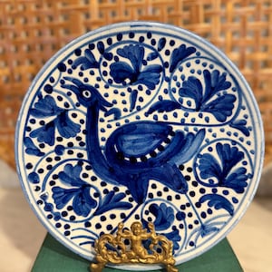 May include: A hand-painted ceramic plate with a blue bird design. The plate features a white background with blue floral and dot patterns. A gold-colored decorative stand supports the plate, which is displayed on a stack of green books.