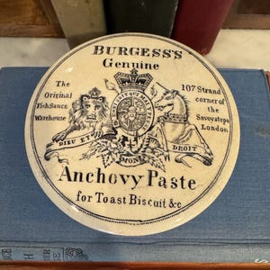 May include: A white ceramic jar lid with black lettering that reads "Burgess's Genuine" and "Anchovy Paste for Toast Biscuit &c". The lid also features a royal crest with a lion and a unicorn, and the text "107 Strand corner of the Savoy steps London".