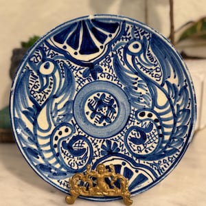 May include: A decorative ceramic plate with a blue and white floral design. The plate is supported by a gold-colored metal stand. The plate's design features intricate patterns and shapes, creating a visually appealing aesthetic. The plate is a round shape.