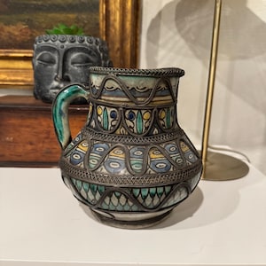 May include: A decorative ceramic pitcher with a green handle and intricate metalwork. The pitcher features a colorful design with blue, yellow, and green accents. The pitcher is a unique piece of home decor.
