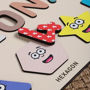 Może przedstawiać: Drewniana zabawka edukacyjna z kolorowymi kształtami i liczbami. Kształty mają twarze, a liczby mają wz&oacute;r strzałek. Tekst "HEXAGON" jest nadrukowany na tablicy.