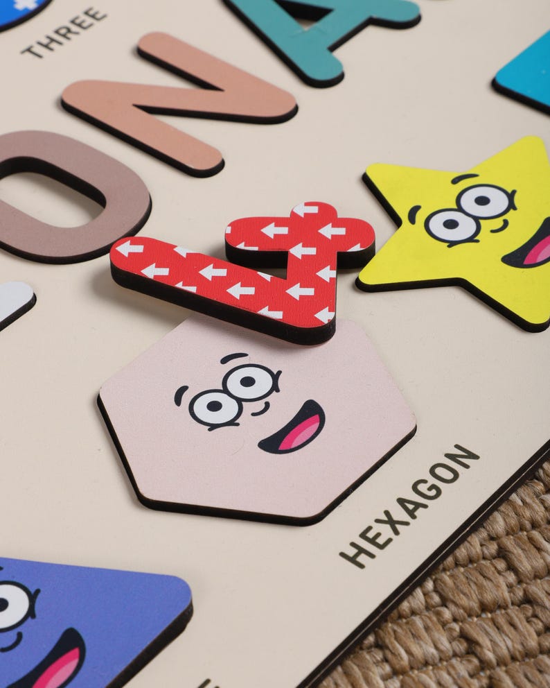 Może przedstawiać: Drewniana zabawka edukacyjna z kolorowymi kształtami i liczbami. Kształty mają twarze, a liczby mają wz&oacute;r strzałek. Tekst "HEXAGON" jest nadrukowany na tablicy.