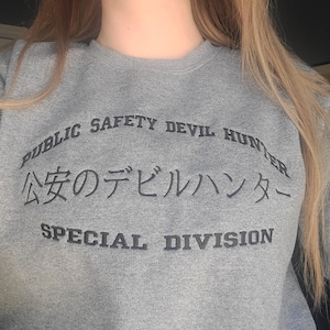 Könnte beinhalten: Grauer Pullover mit dem Text "Public Safety Devil Hunter" und "公安のデビルハンター" in Japanisch sowie "Special Division" darunter.