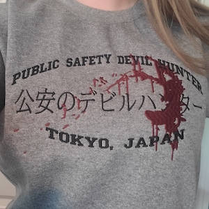 Könnte beinhalten: Grauer Sweatshirt mit dem Text "Public Safety Devil Hunter" und "公安のデビルハンター" in Japanisch. Der Text ist schwarz und es gibt ein rotes Spritzmuster um den Text herum. Der Text "Tokyo, Japan" ist ebenfalls auf dem Sweatshirt aufgedruckt.