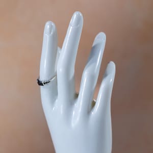 May include: A silver ring featuring a rectangular black stone set in the center, flanked by small black accent stones on each side of the band. The ring is displayed on a white mannequin hand, showcasing the metallic band and the dark stone setting.
