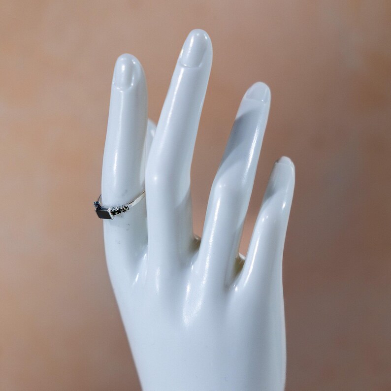 May include: A silver ring featuring a rectangular black stone set in the center, flanked by small black accent stones on each side of the band. The ring is displayed on a white mannequin hand, showcasing the metallic band and the dark stone setting.