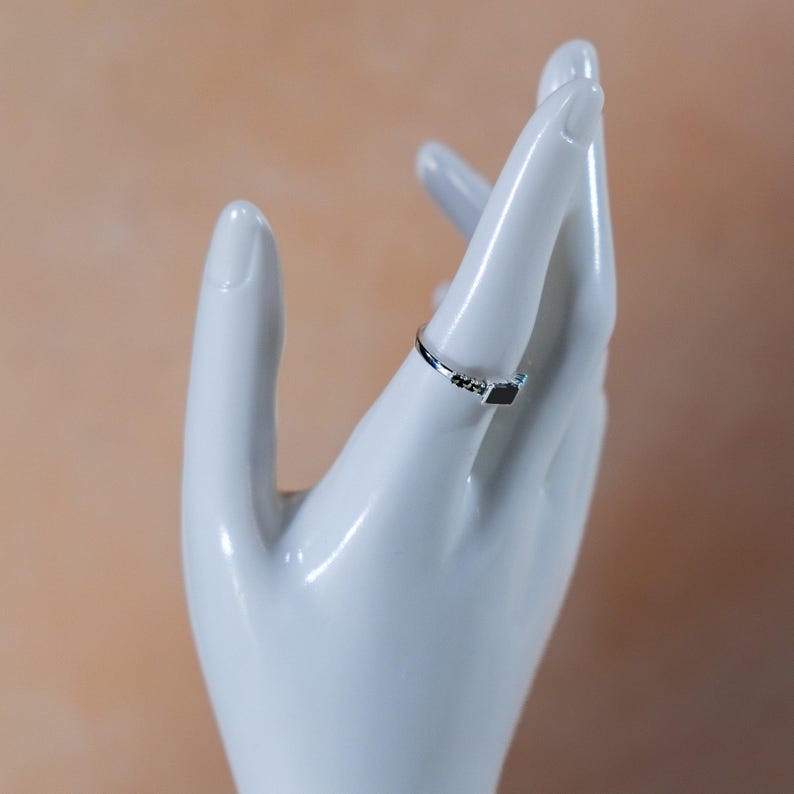 May include: A thin silver-toned ring featuring a small, rectangular black stone set horizontally. The band is slender and metallic, showcasing a minimalist design with a single dark, polished accent piece centered on the top of the ring.