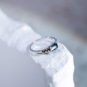 May include: A silver-toned ring featuring a rectangular, dark, iridescent stone set horizontally. The band has a polished finish with small, decorative metallic bead accents flanking the central stone setting on both sides of the ring.