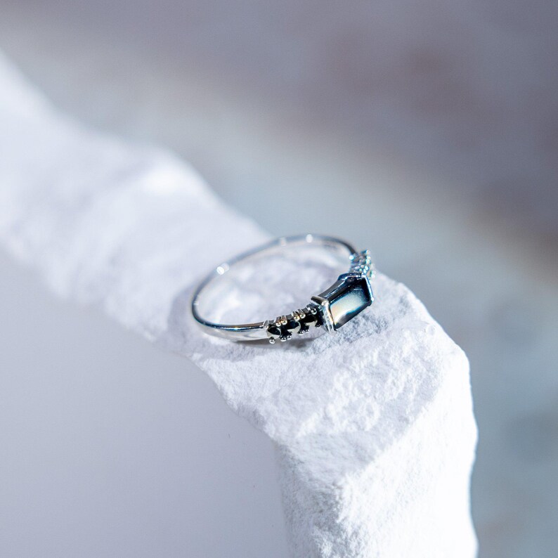 May include: A silver-toned ring featuring a rectangular, dark, iridescent stone set horizontally. The band has a polished finish with small, decorative metallic bead accents flanking the central stone setting on both sides of the ring.
