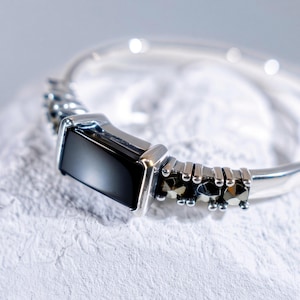 May include: A silver-toned ring featuring a rectangular black stone set in the center. The band is accented with small, dark, faceted stones on either side of the central setting, creating a detailed metallic finish along the ring's shank.