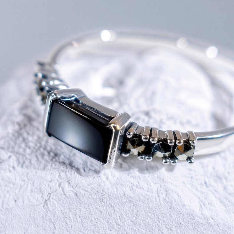 May include: A silver-toned ring featuring a rectangular black stone set in the center. The band is accented with small, dark, faceted stones on either side of the central setting, creating a detailed metallic finish along the ring's shank.