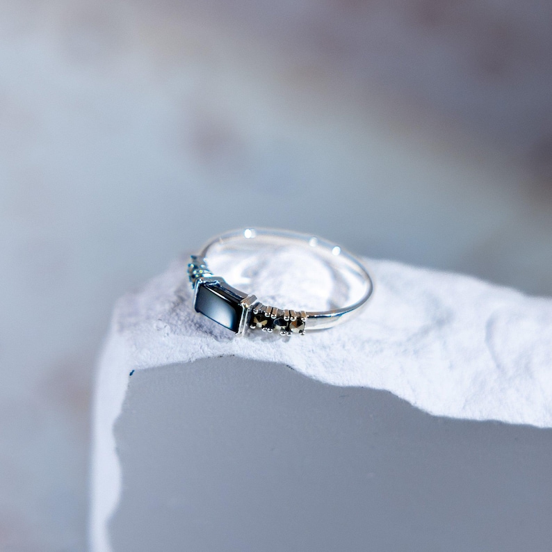 May include: A silver ring featuring a central rectangular black stone flanked by two smaller dark stones on each side. The band is thin and metallic, showcasing a polished finish that reflects light across its surface and the dark gemstone setting.