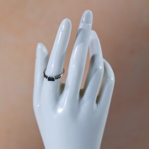 May include: A silver-toned ring featuring a central rectangular black stone set horizontally. The band is thin and metallic, accented with small, dark, bead-like details flanking the central stone on both sides, creating a refined look.