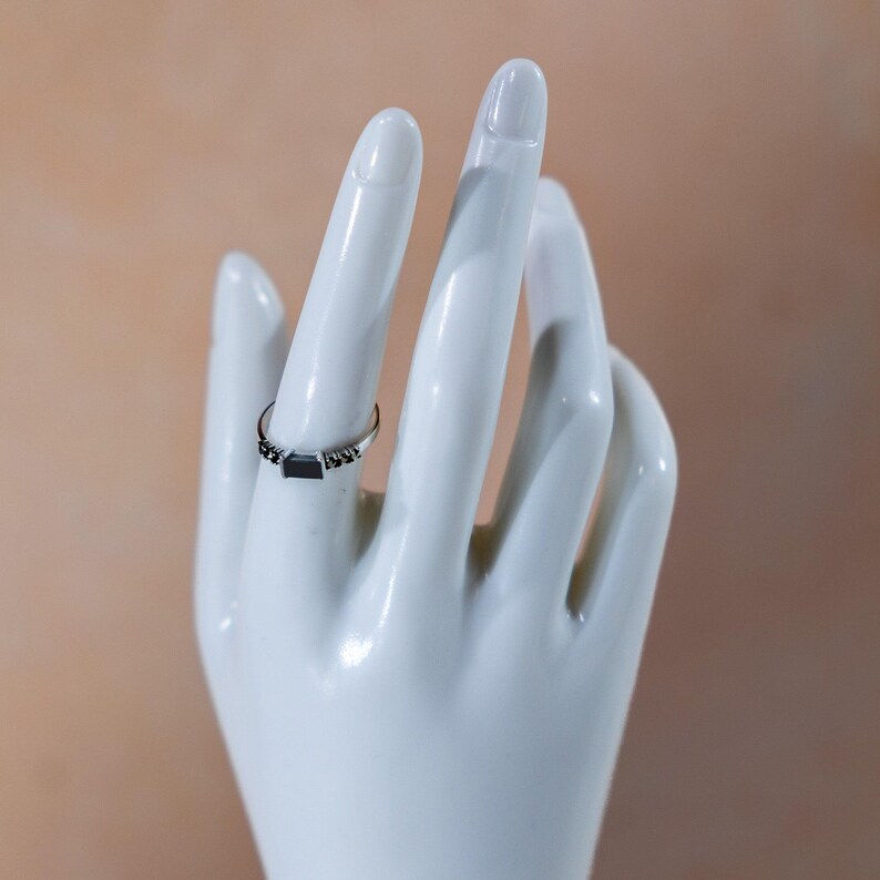 May include: A silver-toned ring featuring a central rectangular black stone set horizontally. The band is thin and metallic, accented with small, dark, bead-like details flanking the central stone on both sides, creating a refined look.