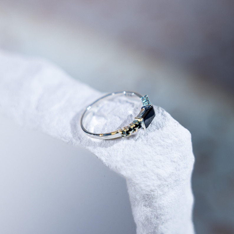 May include: A silver-toned ring featuring a central rectangular black stone flanked by smaller, clear, light-reflecting stones set along the band. The metal band is thin and polished, showcasing a refined design with a prominent dark gemstone centerpiece.