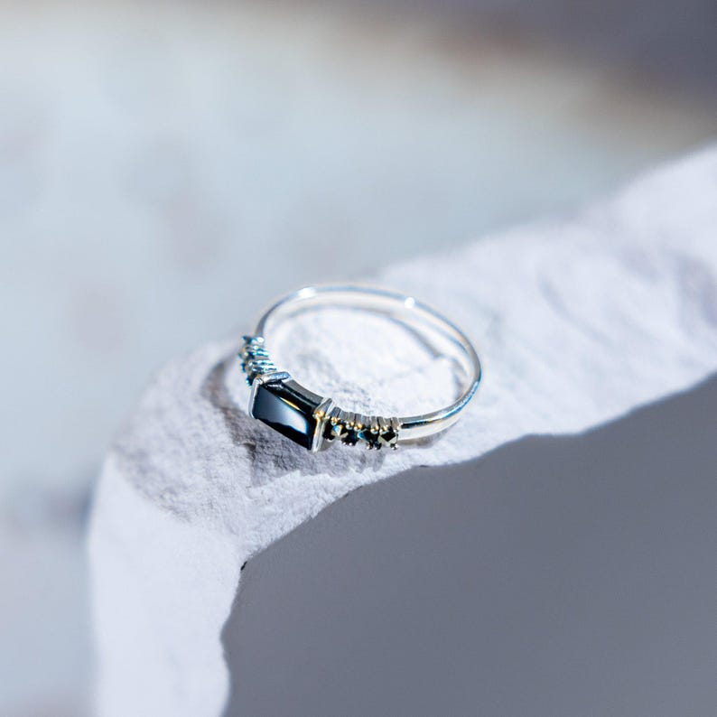 May include: A silver ring featuring a central rectangular black gemstone flanked by three small dark stones on each side. The band is thin and polished, showcasing a sleek design with a prominent dark stone setting that catches the light.