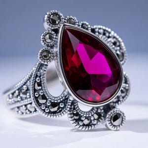 May include: A silver ring featuring a large, pear-shaped, deep red gemstone as the centerpiece. The metal band and setting are decorated with numerous small, dark, faceted stones arranged in a scalloped, ornate pattern surrounding the center.