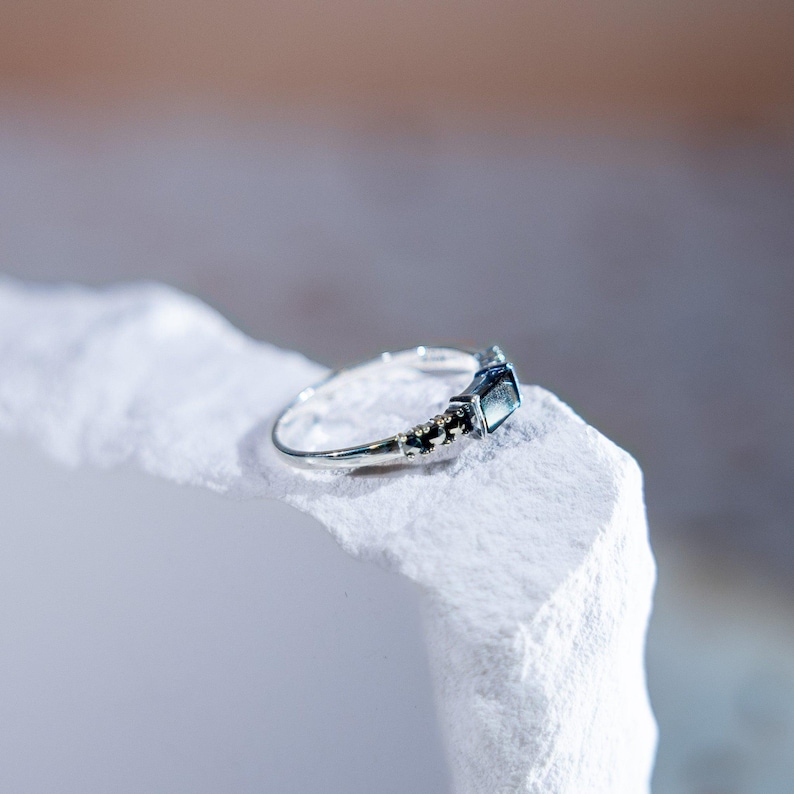 May include: A silver-toned ring featuring a rectangular dark blue gemstone set in the center, flanked by small, dark, faceted stones along the band. The metal band is thin and polished, showcasing a clear, reflective surface on the main stone.