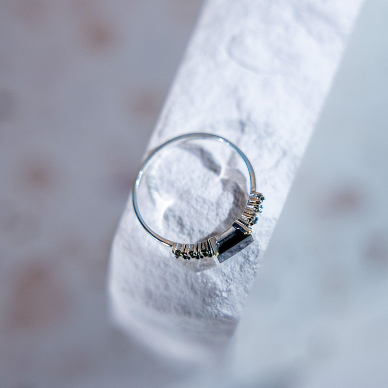 May include: A silver-toned ring featuring a central rectangular dark stone flanked by a series of small, dark, round-cut stones set along the band. The metal band is thin and polished, showcasing a clear contrast between the dark stones and metal.