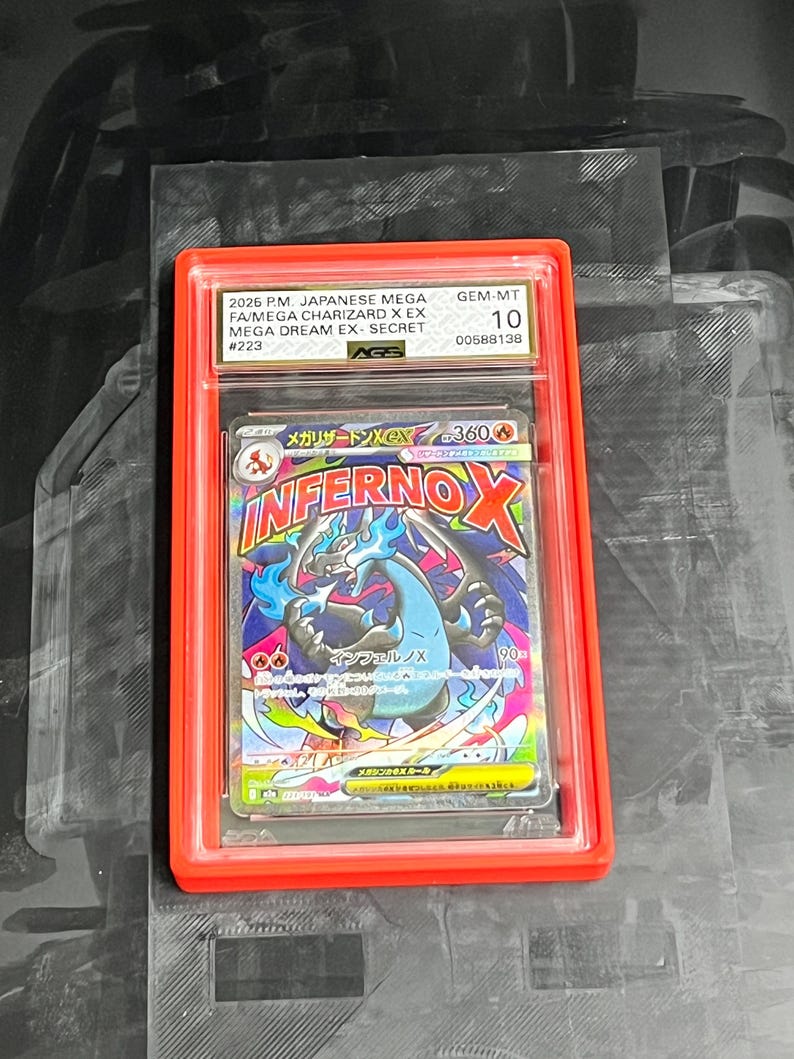 May include: A graded Pok&eacute;mon card featuring a Charizard EX. The card is encased in a red holder with the text "2026 P.M. JAPANESE MEGA FA/MEGA CHARIZARD X EX MEGA DREAM EX SECRET #223" and "GEM-MT 10". The card art displays a blue and black Charizard with the text "INFERNO X".