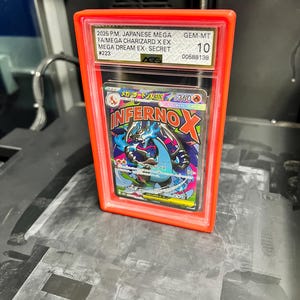 May include: A graded trading card in a clear plastic case with an orange frame. The card features a colorful illustration of a dragon-like creature with the text "INFERNO X". The case has text indicating it is a Japanese Mega Charizard card.