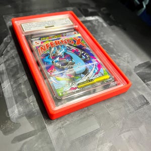 May include: A graded trading card in a red plastic case. The card features a colorful illustration of a blue and black creature with the word "InfernoX" in red and yellow. The card is graded a 10.