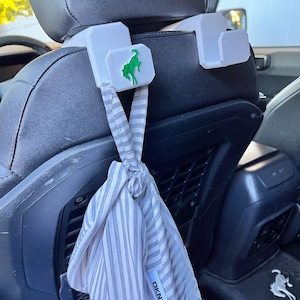 May include: White car seat headrest hooks with a green horse logo. A gray and white striped DKNY scarf is tied to the hooks. The hooks are attached to a black car seat.