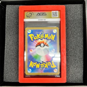 May include: A graded Pok&eacute;mon trading card in a red plastic holder. The card is visible through a clear plastic case. The holder has the AGS logo and a QR code. The card has a blue background with the Pok&eacute;mon logo.