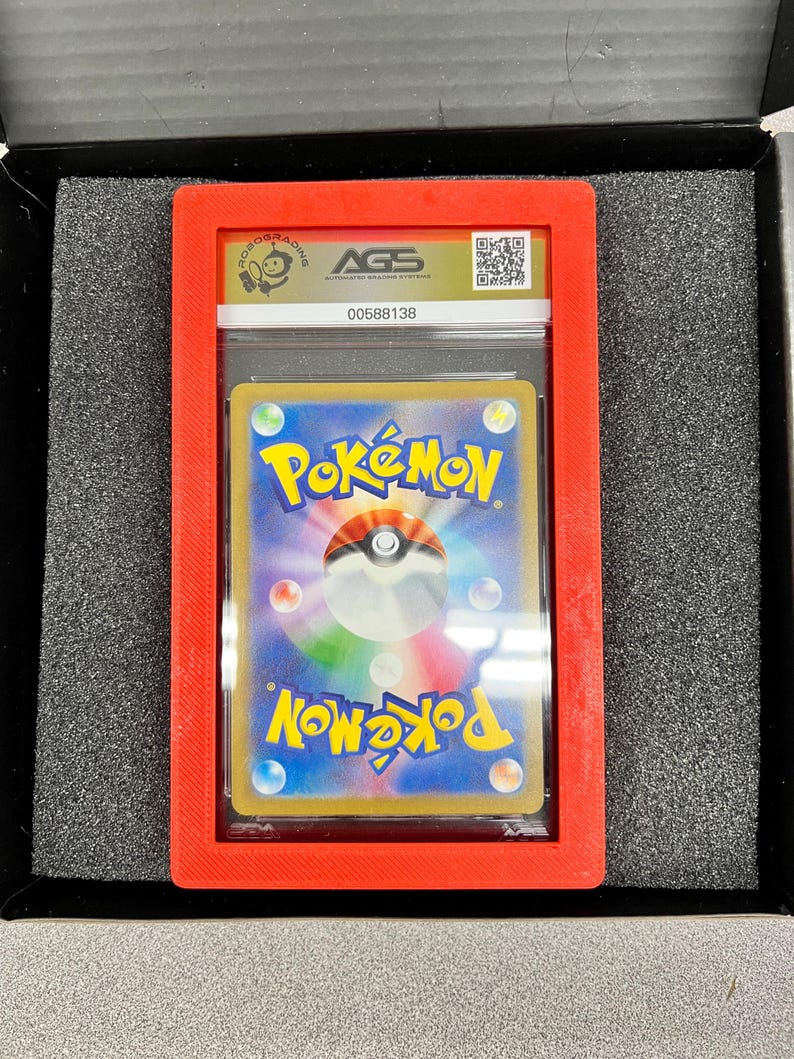 May include: A graded Pok&eacute;mon trading card in a red plastic holder. The card is visible through a clear plastic case. The holder has the AGS logo and a QR code. The card has a blue background with the Pok&eacute;mon logo.