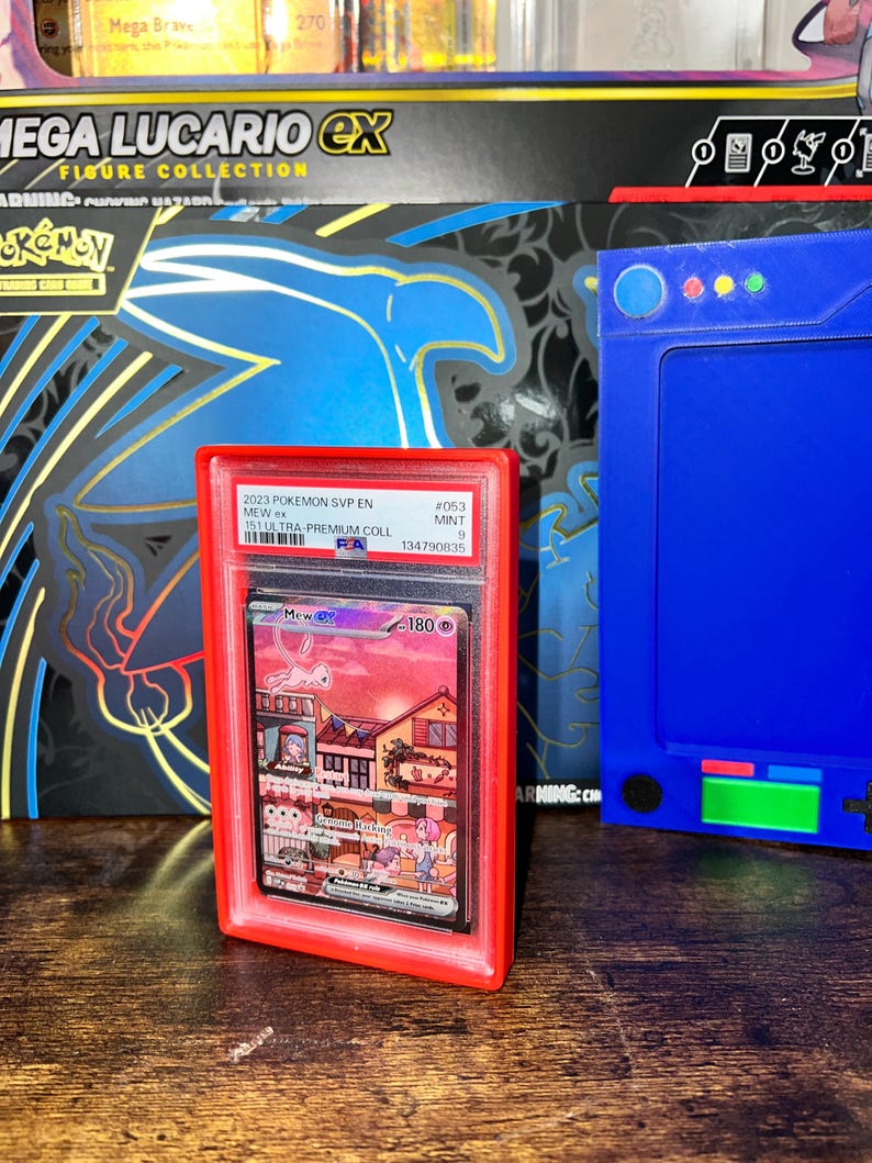 May include: A graded Pok&eacute;mon card featuring Mew ex, encased in a red holder. The card displays a colorful illustration of Mew and a town scene. The holder is labeled with the card's details, including the set and grade.