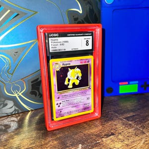 May include: A graded Pok&eacute;mon card of Hypno from the Fossil set, encased in a red protective holder. The card is graded NM/MINT 8 by CGC. The card features a yellow and purple design with the name "Hypno" and the HP value.