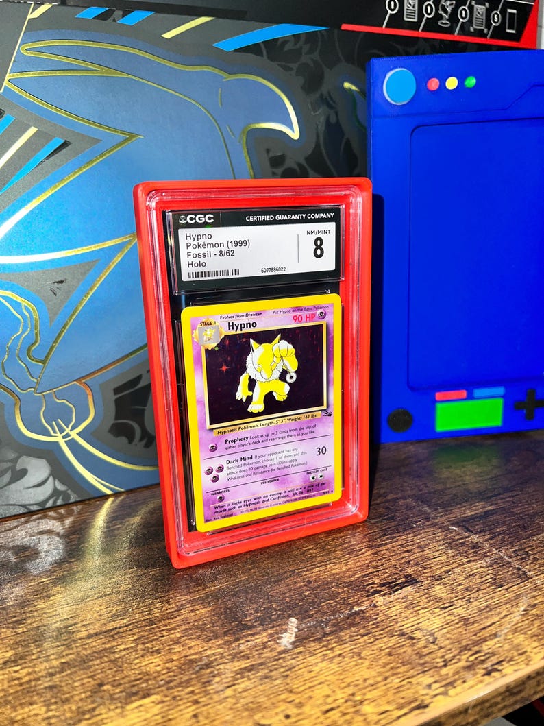May include: A graded Pok&eacute;mon card of Hypno from the Fossil set, encased in a red protective holder. The card is graded NM/MINT 8 by CGC. The card features a yellow and purple design with the name "Hypno" and the HP value.