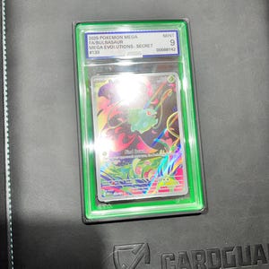 May include: A graded Pok&eacute;mon card featuring a Mega Bulbasaur from the Mega Evolutions Secret set. The card is encased in a green holder with the text "MINT 9" and other details. The card art showcases a colorful illustration of Bulbasaur.