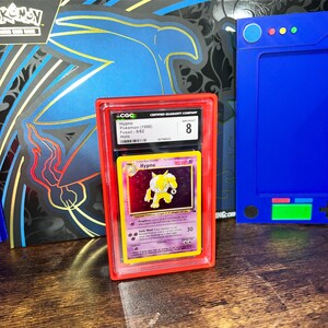 May include: A graded Pok&eacute;mon card featuring Hypno from the Fossil set, encased in a red protective holder. The card is graded an 8 by CGC. The background includes a blue and black box with the text "MEGA LUCARIO EX FIGURE COLLECTION".
