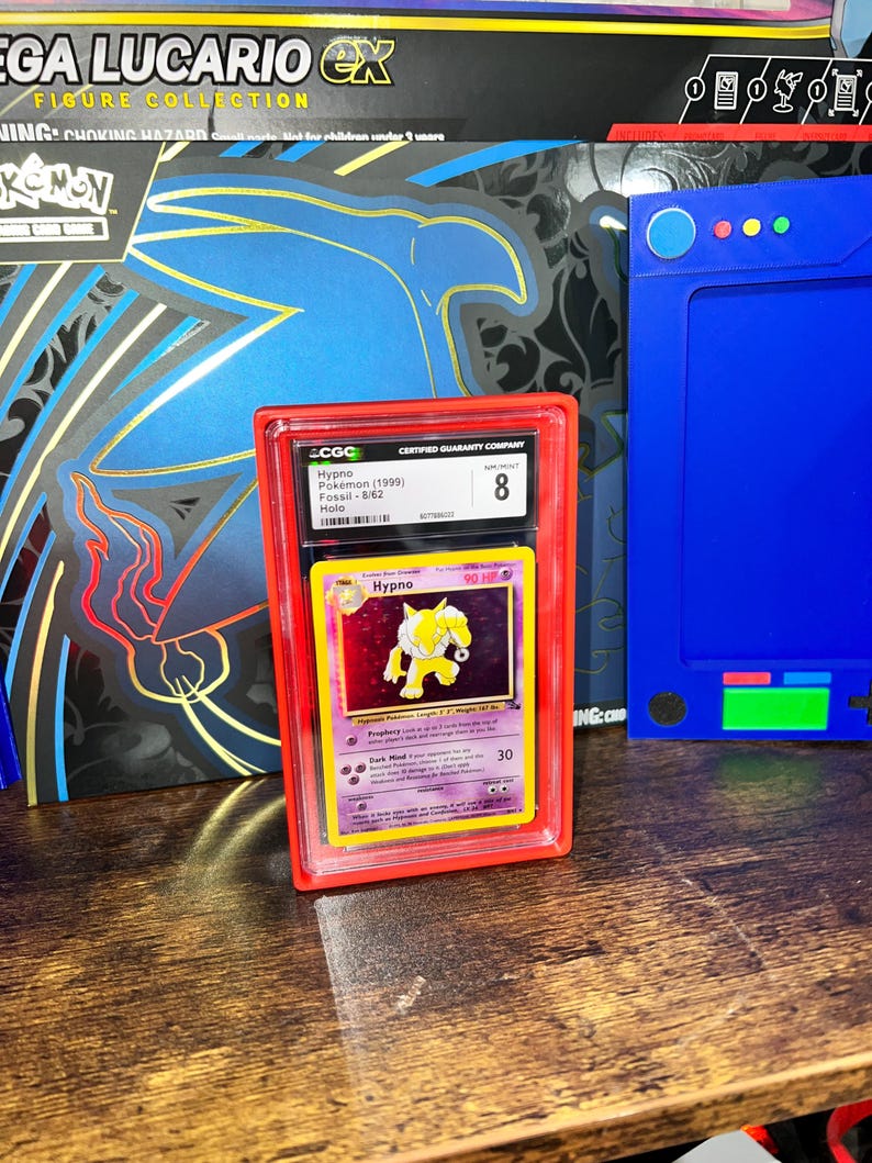May include: A graded Pok&eacute;mon card featuring Hypno from the Fossil set, encased in a red protective holder. The card is graded an 8 by CGC. The background includes a blue and black box with the text "MEGA LUCARIO EX FIGURE COLLECTION".
