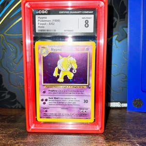 May include: A graded Hypno Pok&eacute;mon card from the Fossil set, 8/62, in a red protective case. The card features a yellow Hypno character on a purple background. The card is graded NM/MINT 8 by CGC.