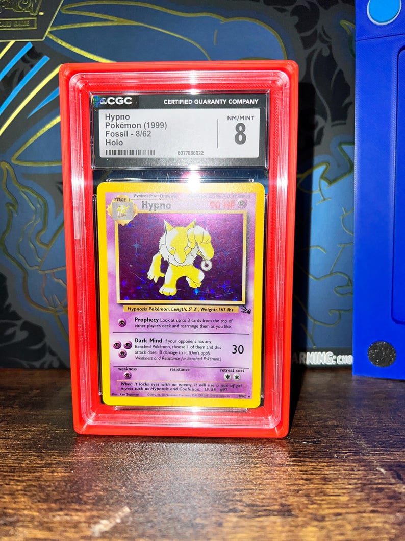 May include: A graded Hypno Pok&eacute;mon card from the Fossil set, 8/62, in a red protective case. The card features a yellow Hypno character on a purple background. The card is graded NM/MINT 8 by CGC.