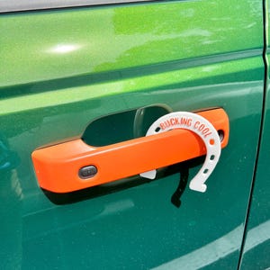 May include: An orange car door handle with a white horseshoe-shaped accessory. The accessory has the words "BUCKING COOL" printed on it. The car door is green.