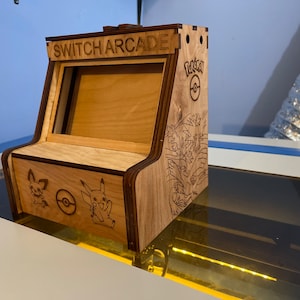 以下が含まれることがあります： ポケモンをテーマにした木製ニンテンドースイッチアーケードスタンド。スタンドはスイッチの画面用の切り抜きがあり、側面にポケモンのロゴとキャラクターが描かれています。上部には「SWITCH ARCADE」という文字が刻まれています。