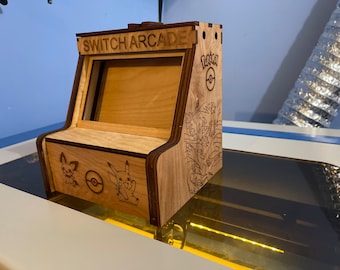 May include: A wooden Nintendo Switch arcade stand with a Pokemon theme. The stand has a cutout for the Switch screen and features a Pokemon logo and characters on the side. The text "SWITCH ARCADE" is engraved on the top.