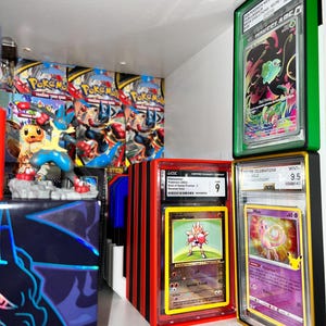 May include: A display of graded Pok&eacute;mon trading cards in protective cases. The cases are various colors, including green, red, and black. The cards feature images of Pok&eacute;mon characters like Hitmonchan and Mew. Background includes Pok&eacute;mon booster packs and a Lucario figurine.